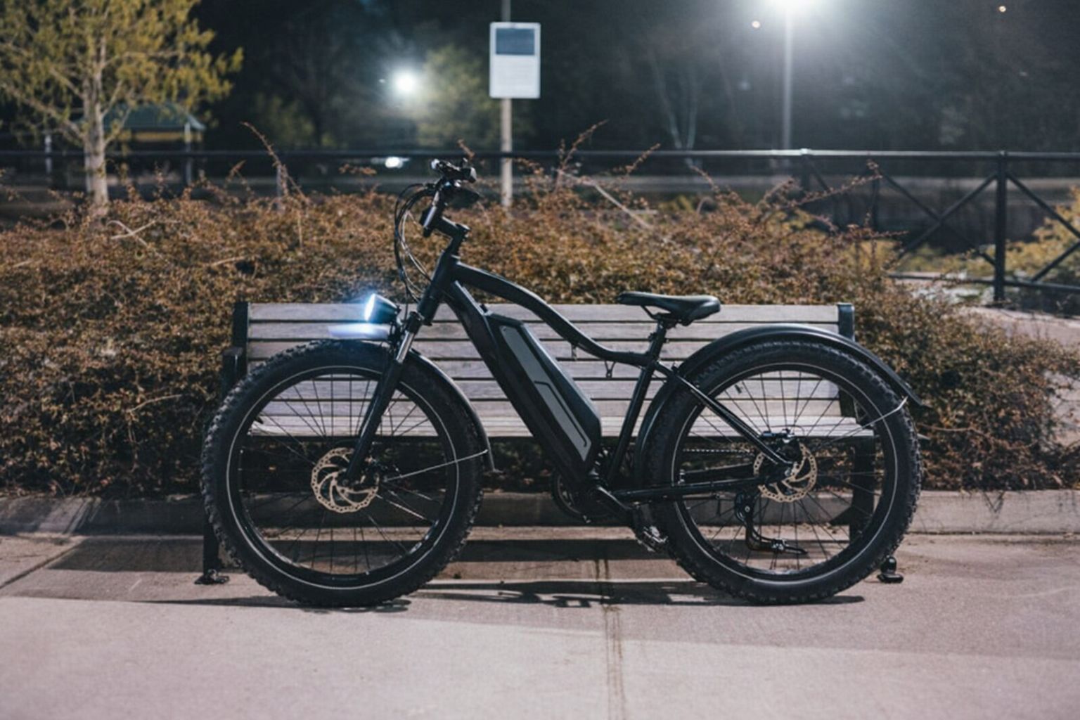 Svart elcykel med breda däck parkerad framför en parkbänk på kvällen.